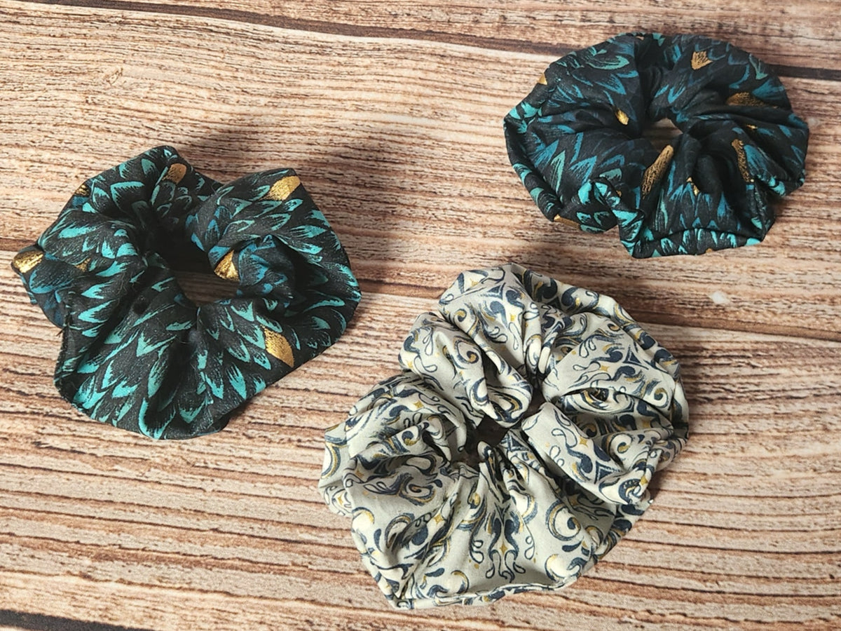 Three handmade Dragon collection scrunchies shown separately: two teal and black dragon-scale scrunchies with metallic gold accents and one light dragon-themed scrunchie.