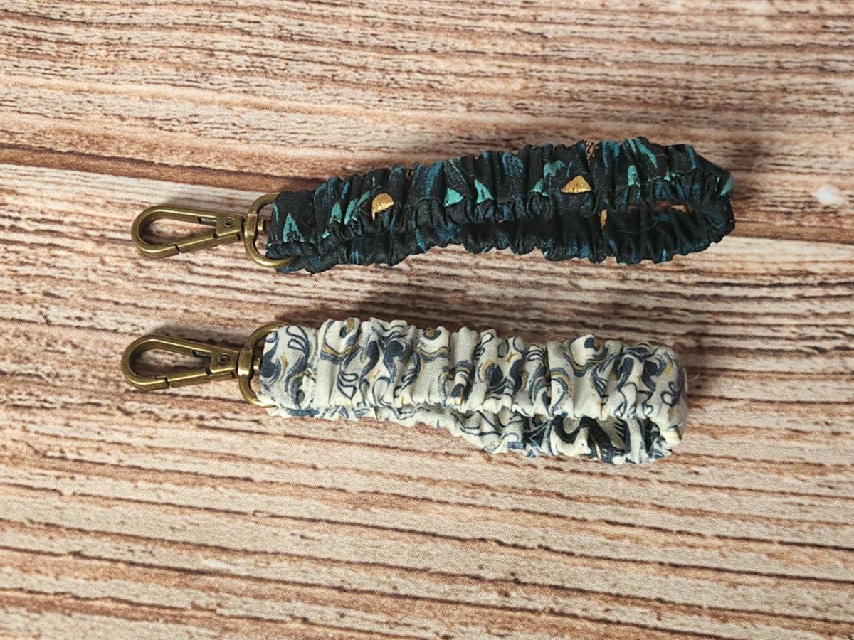 Two handmade scrunchie keychains from the Dragon collection, one in teal and black dragon-scale fabric with metallic gold accents and one in a light dragon-themed print, each with antique-brass-toned swivel hardware.