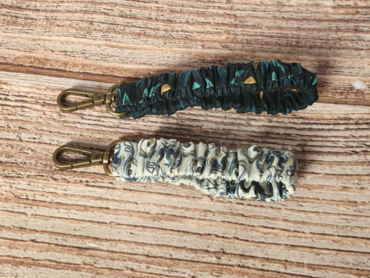 Two handmade scrunchie keychains from the Dragon collection, one in teal and black dragon-scale fabric with metallic gold accents and one in a light dragon-themed print, each with antique-brass-toned swivel hardware.