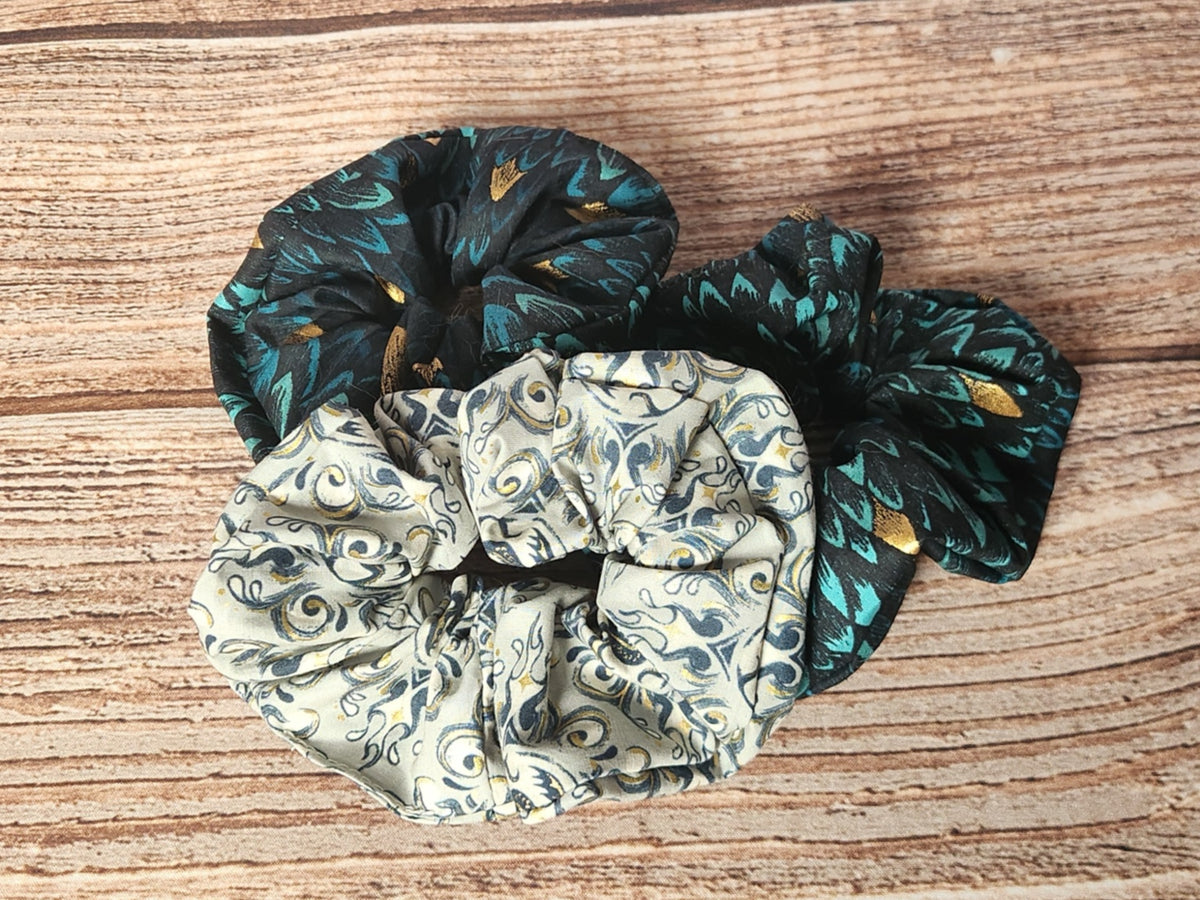 Three handmade scrunchies from the Dragon collection: two in teal and black dragon-scale fabric with metallic gold accents and one in a light coordinating dragon-themed print.