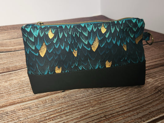 Front view of a handmade angled zipper pouch with teal and black dragon-scale fabric, metallic gold accents, a solid black lower panel, antique-brass zipper hardware, and a side D-ring.