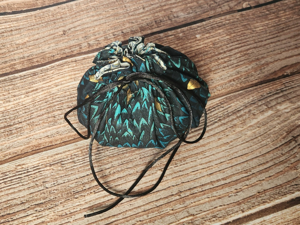 Closed handmade drawstring dice bag in teal and black dragon-scale fabric with metallic gold accents, black drawcords, and a light coordinating ruffled lining at the opening.