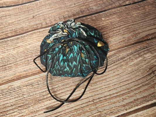 Closed handmade drawstring dice bag in teal and black dragon-scale fabric with metallic gold accents, black drawcords, and a light coordinating ruffled lining at the opening.