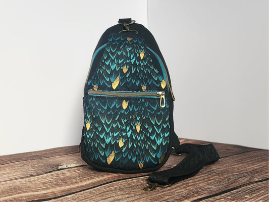 Front view of a handmade sling bag with teal and black dragon-scale fabric, metallic gold accents, black side panels, an antique-brass front zipper, and a black adjustable strap.